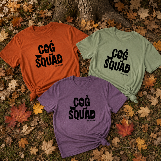 Three graphic T-shirts in orange, sage green, and heather purple are arranged in a flat lay on a bed of fallen autumn leaves and tree bark. Each shirt features a black 'COG SQUAD' graphic with a small bird icon above the text.